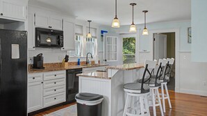 A kitchen with white cabinetry, a black refrigerator, a microwave, and a coffee maker. There are three bar stools and a trash can. - 321 Williston Rd (Bourne)