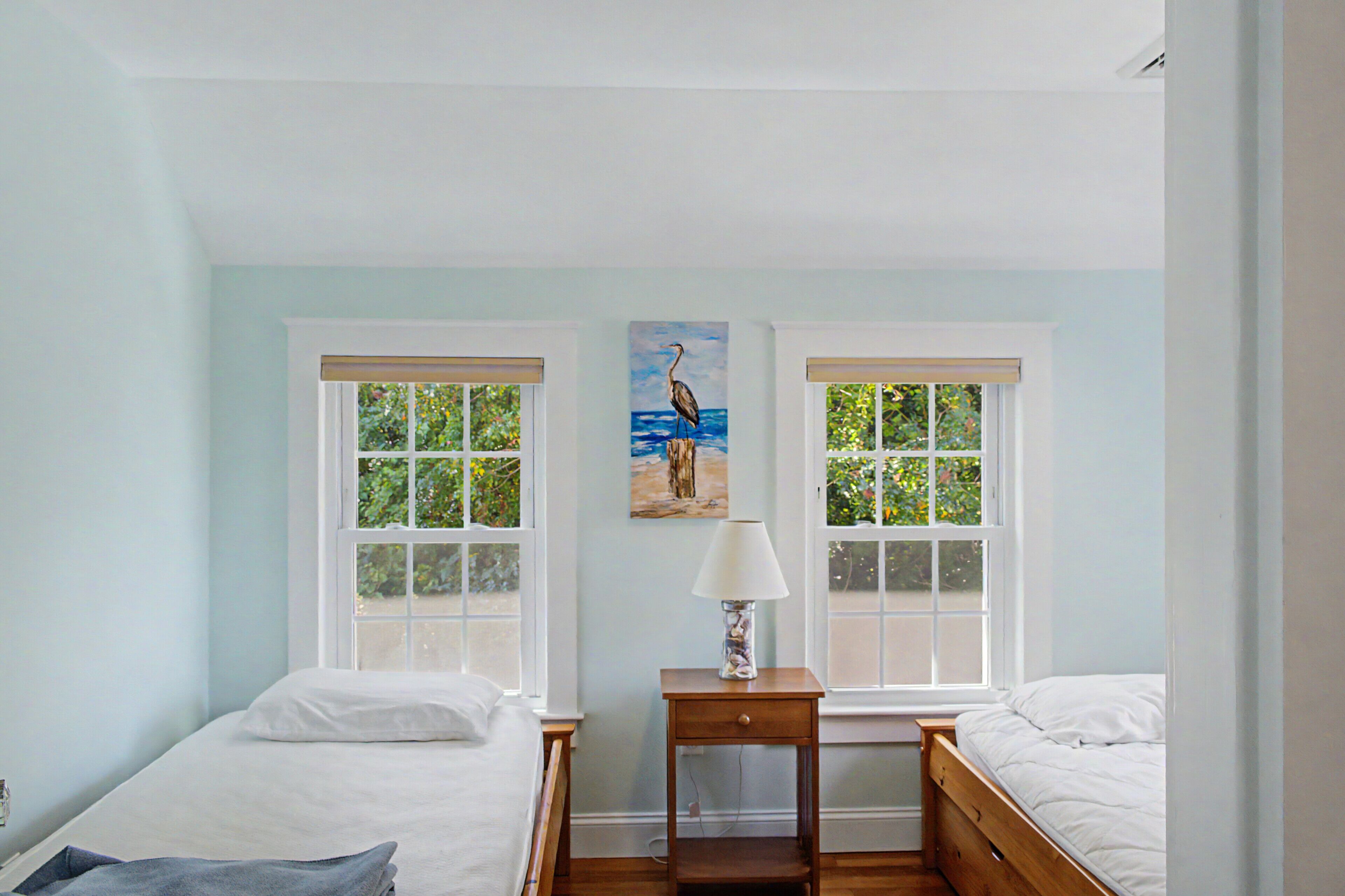 A room with two beds, a nightstand, a painting, and windows with a view of greenery.