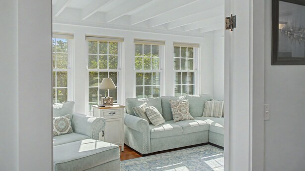A room with a light blue sofa, a small table with a lamp, and large windows. - 321 Williston Rd (Bourne)
