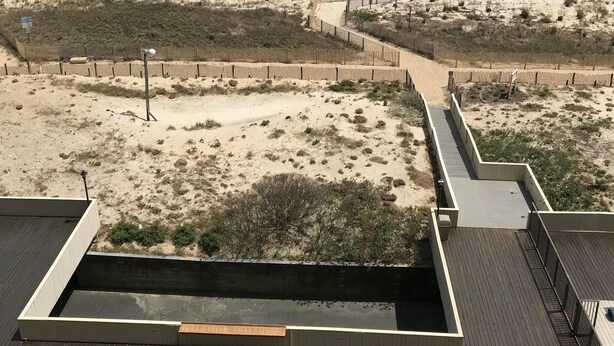 En vy över stranden från en högt belägen plats, med en sandstrand, havet och en träbrygga med en inbyggd sittplats.