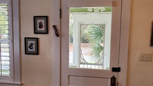 Een witte deur met een decoratief glasraam, een klein tafeltje met een gestreept kussen, en twee ingelijste foto's aan de muur.