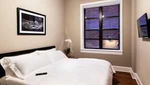 A neatly made bed with white linens, a framed picture on the wall, a window with a brick pattern, and a television mounted on the wall.