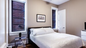 A bedroom with a bed, a window with a view of a brick wall, a ceiling fan, and a framed picture on the wall.