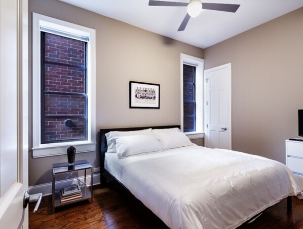 A bedroom with a bed, a window with a view of a brick wall, a ceiling fan, and a framed picture on the wall.