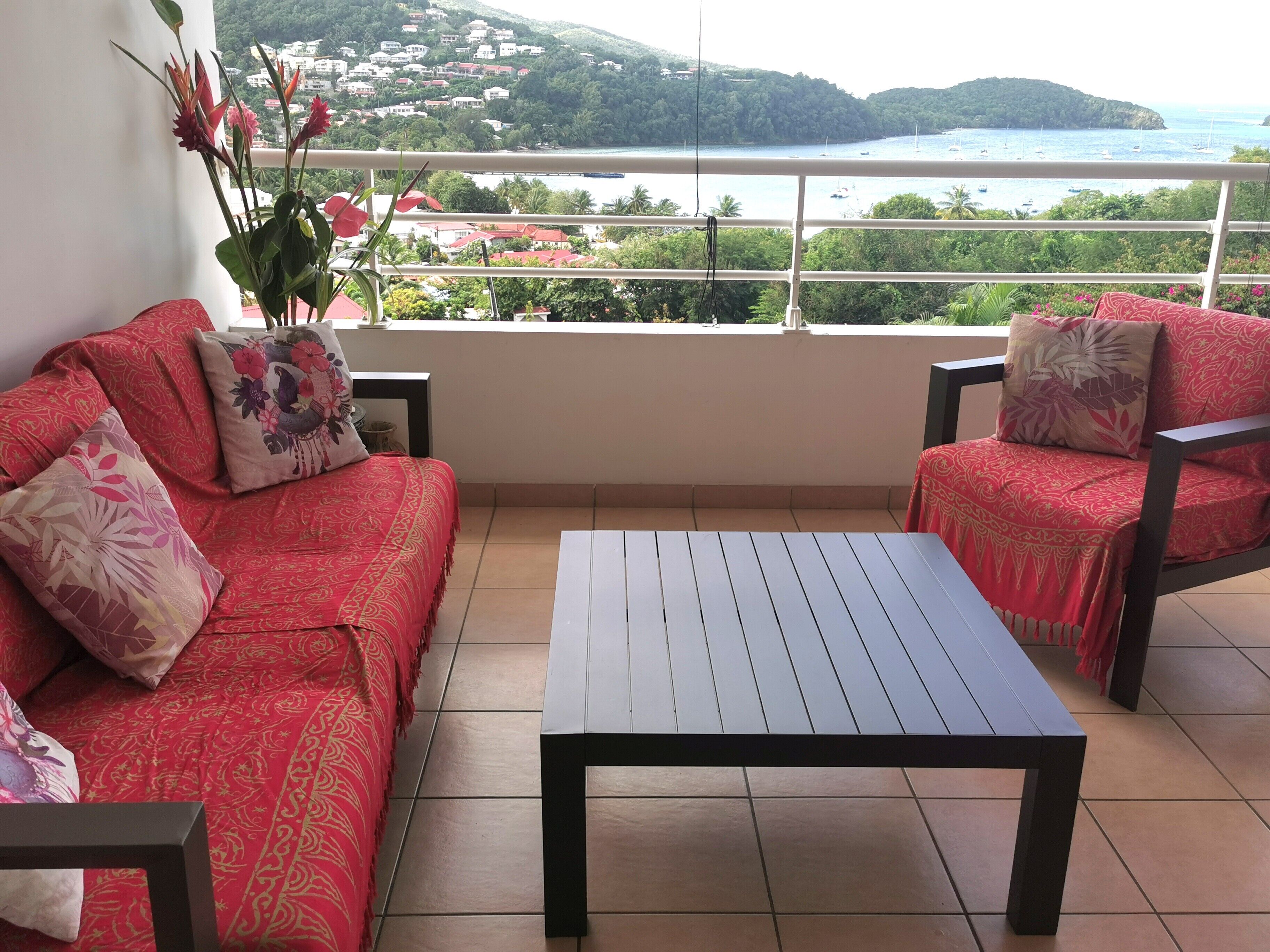 A balcony with a red sofa and two chairs, a white table, and a view of a coastal area with boats and houses.