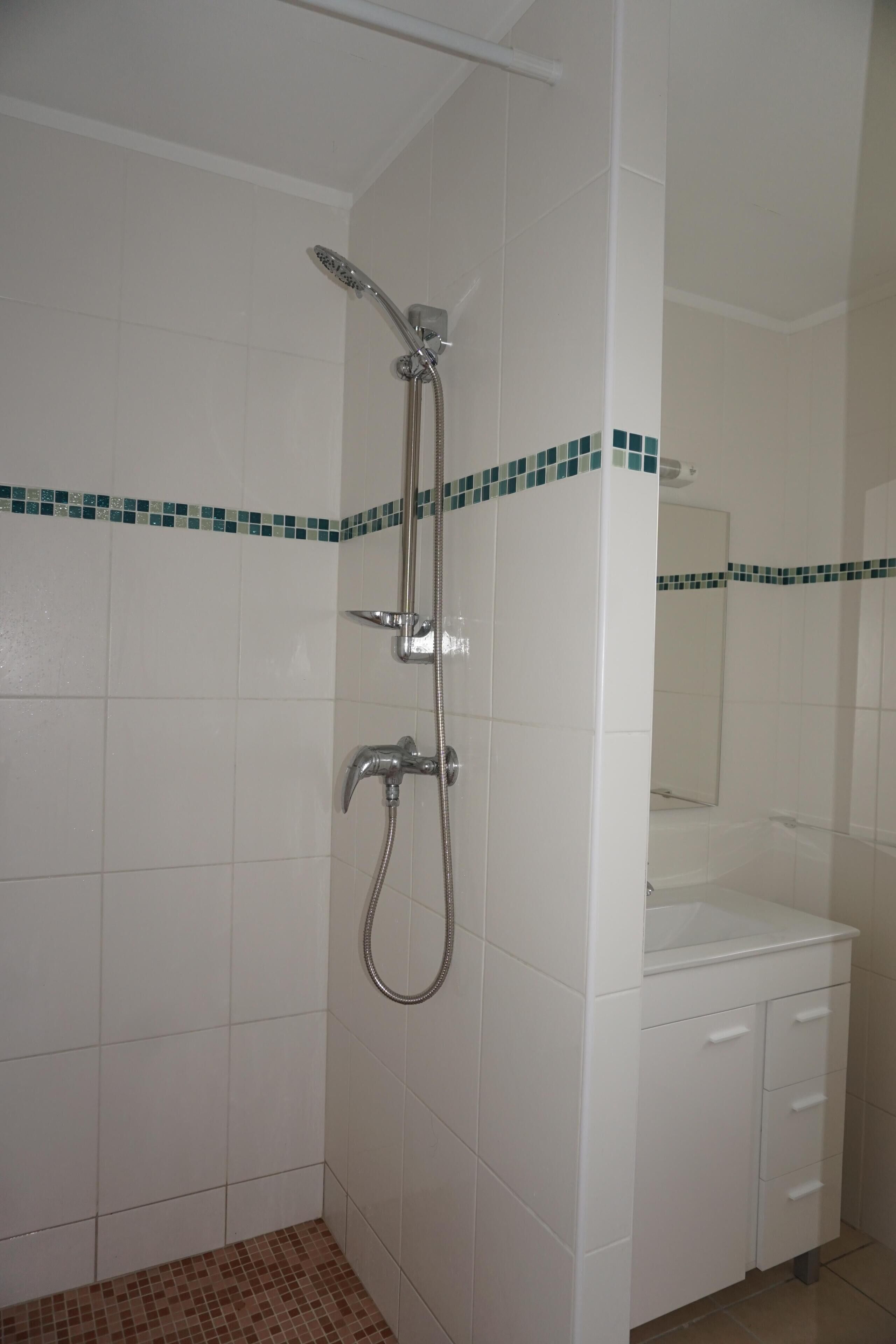 Une salle de bain avec des carreaux blancs, une douche avec un pommeau de douche amovible, un robinet mural et un petit lavabo avec un miroir.