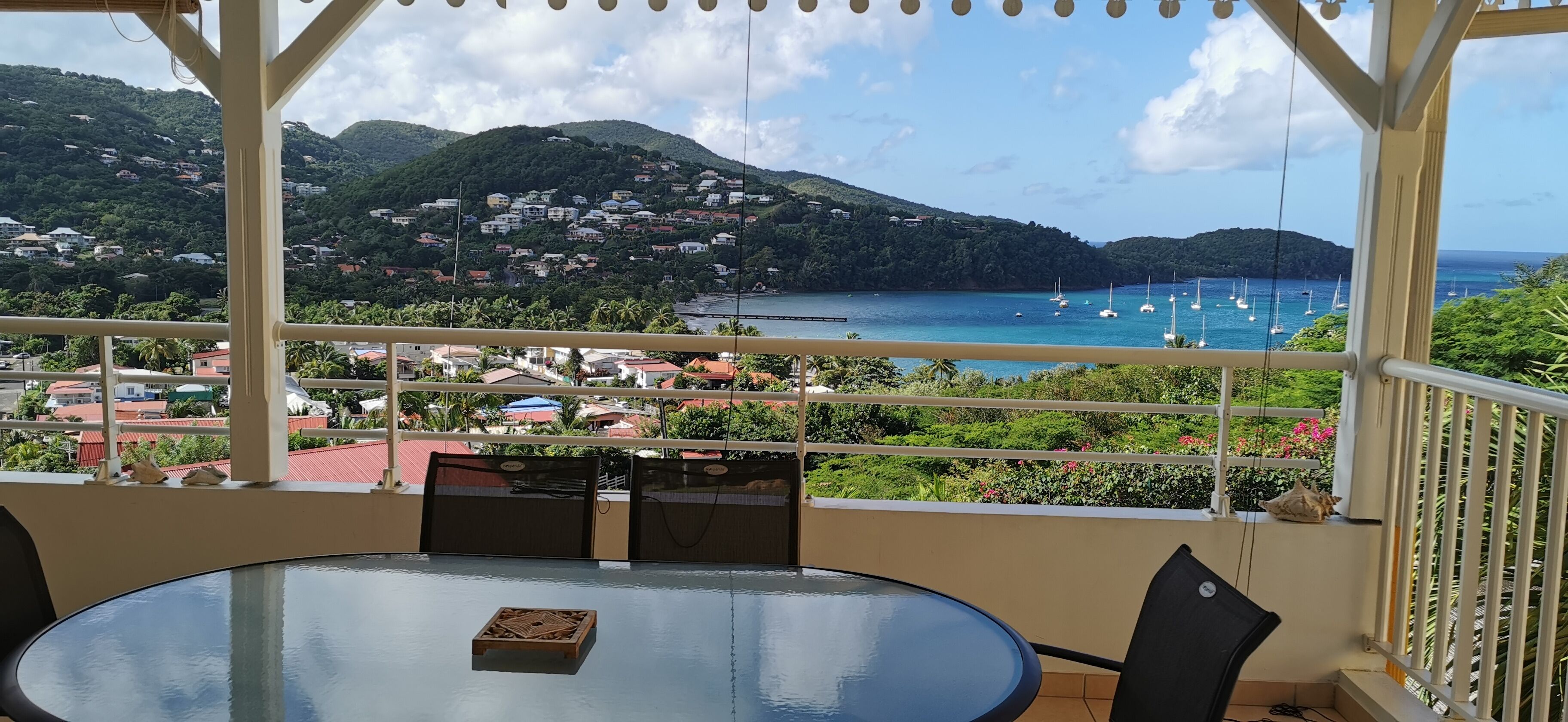 A balcony with a table and chairs, overlooking a coastal area with houses, greenery, and boats.