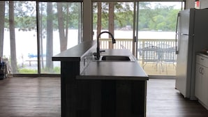 Een keuken met een centraal eiland, een spoelbak en een koelkast. Een groot raam biedt uitzicht op bomen en een wateroppervlak.