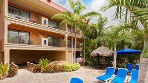 A hotel with a two-story building, outdoor lounge area with blue chairs, palm trees, and a thatched umbrella. - Coconut Cove All-suite (Clearwater Beach)