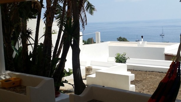 Una hamaca bajo un techo de madera, con vistas a un edificio blanco y palmeras, y vista al mar.