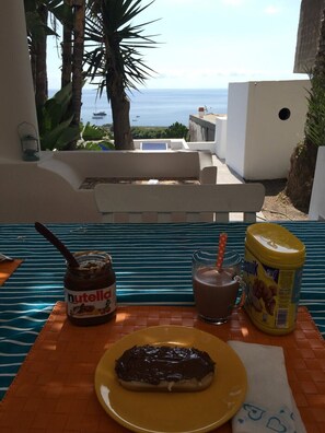 A table with a Nutella jar, a glass of chocolate milk, and a chocolate-covered pastry on a yellow plate, set against a backdrop of a balcony with a view of the sea and palm trees. - Stromboli, At Nino's House (Stromboli)