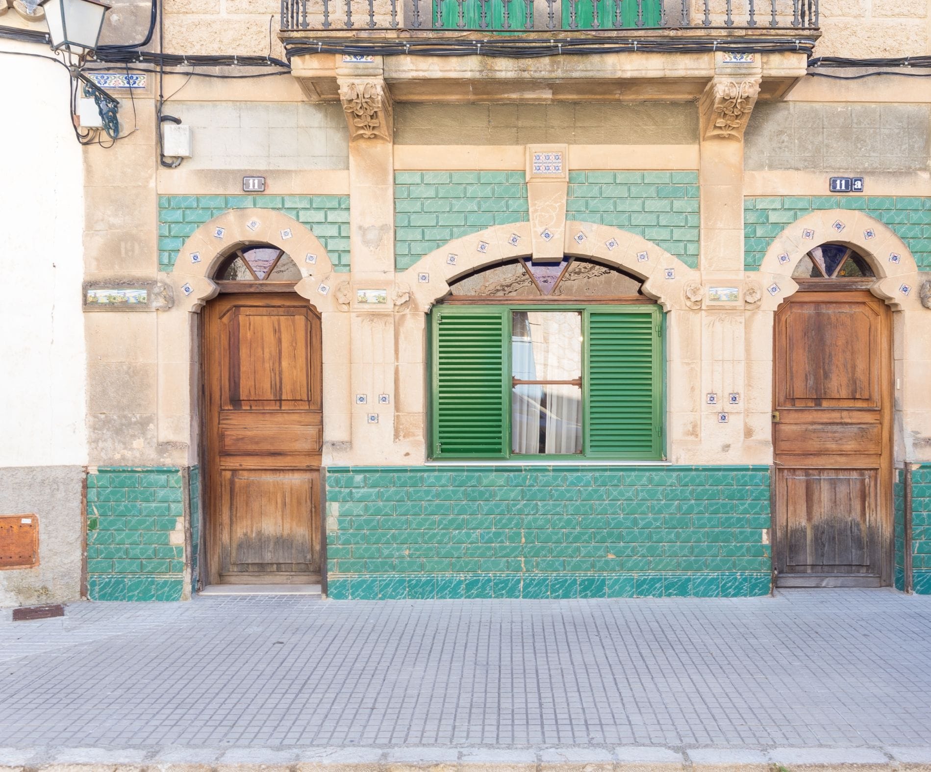 A building facade with two wooden doors, a window with green shutters, and a tiled wall with green and beige patterns.