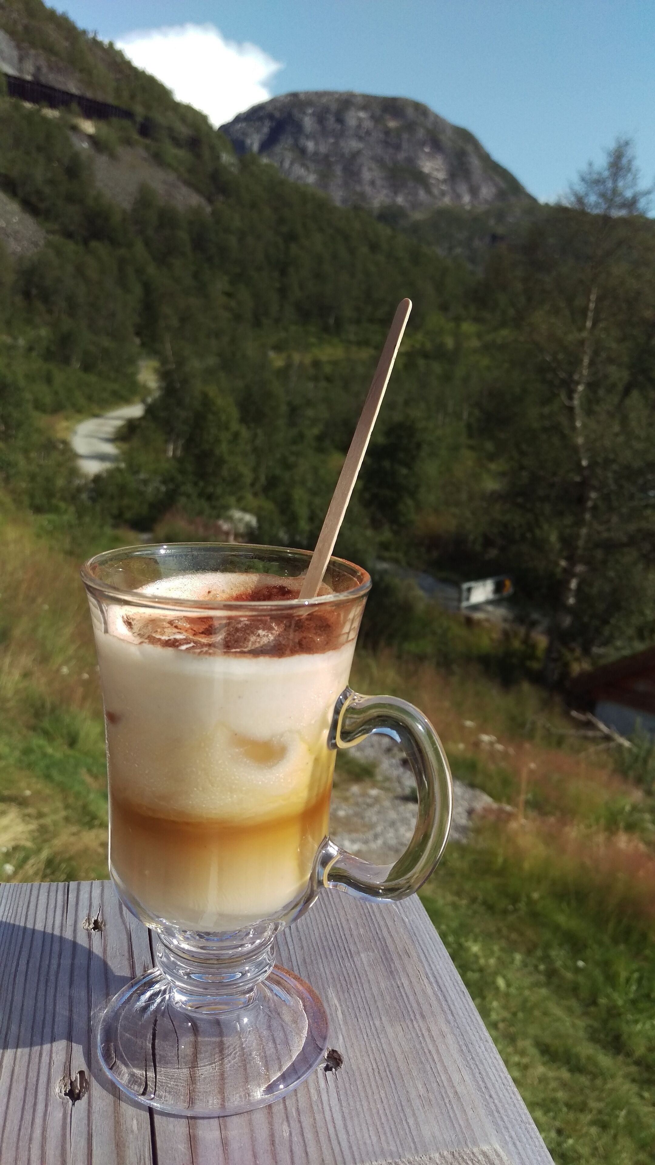 Un vaso con una bebida en capas y una pajita, sobre una superficie de madera, con un paisaje montañoso de fondo.