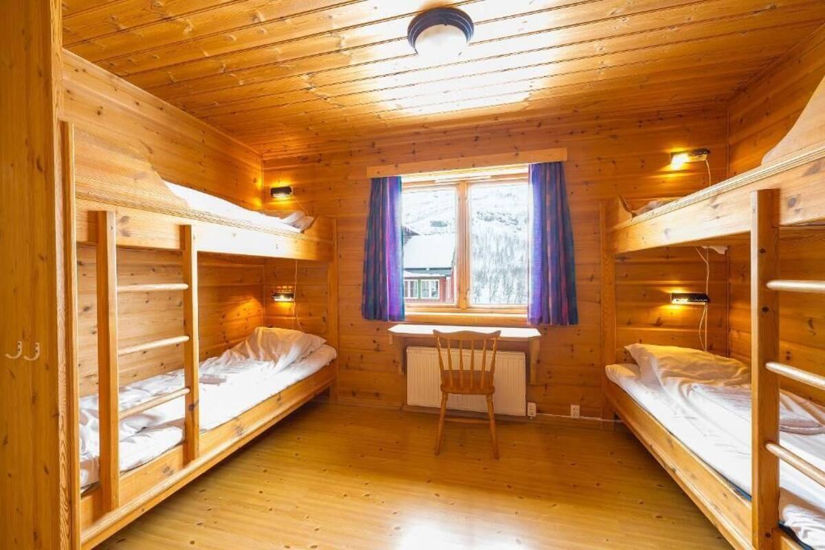 Une cabine aux murs et au plancher en bois, avec deux lits superposés, un bureau avec une chaise et une fenêtre avec des rideaux.