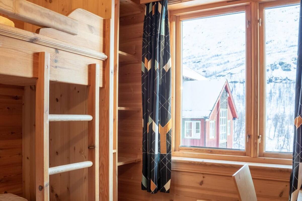 Une chambre dans un chalet en bois, avec un lit superposé, une fenêtre avec des rideaux, un radiateur et une chaise.