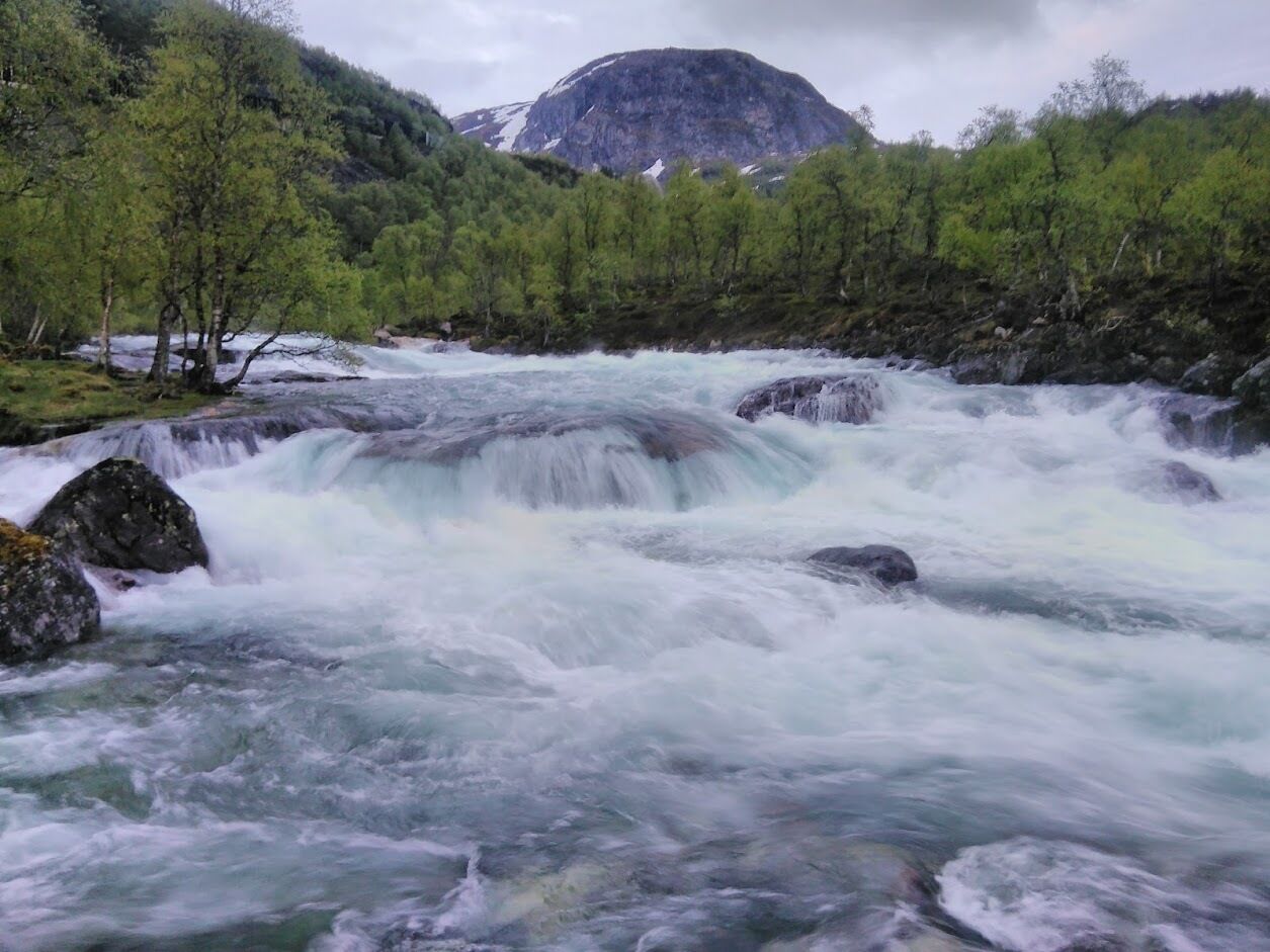A river with a waterfall, surrounded by green trees and a mountain in the background.