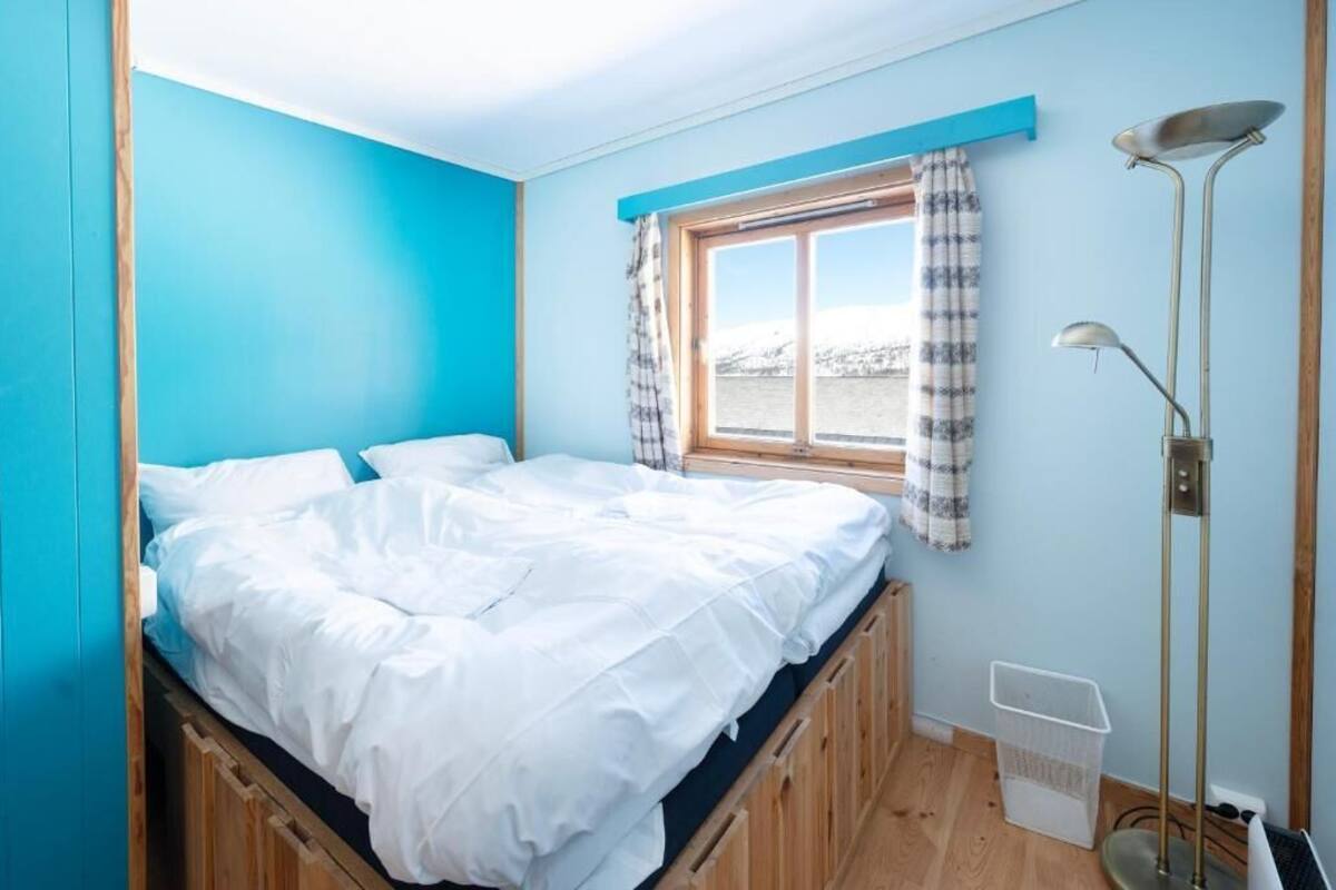 Une pièce avec un lit, une fenêtre avec des rideaux, une lampe sur pied et une poubelle.