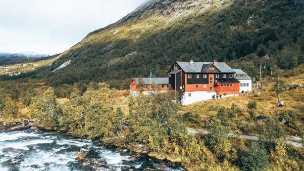 Un edificio grande de varios pisos con fachada roja y blanca, rodeado de un bosque denso y un río.
