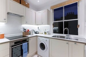 A modern kitchen with white cabinetry, a built-in oven, a washing machine, and a window with a city view at night. - Attractive Chelsea apartment for four (London)