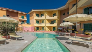 A pool area with lounge chairs and umbrellas, surrounded by a multi-story building with balconies. - One bedroom apartment (4 people) - Residence I Mirti Bianchi (Santa Teresa Gallura)