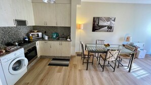 A compact kitchen with a microwave, stove, and washing machine. A dining table with chairs is in the center, and a painting hangs on the wall. - Apartment/ flat - Coruña (O Pindo)