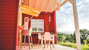 Un bâtiment rouge doté d’un patio abrité, avec une table et des chaises, une enseigne indiquant « TI-PUNCH BAR » et une vue sur la verdure en arrière-plan.