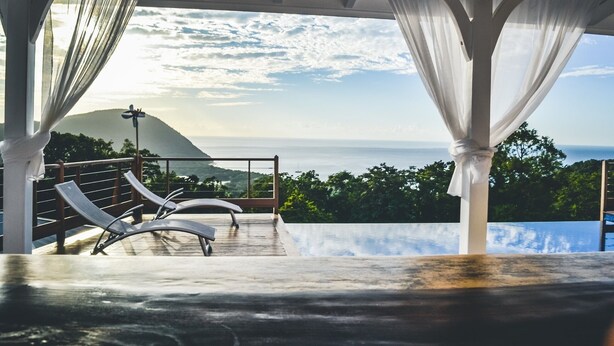 Un espace extérieur aménagé avec une zone salon couverte, des rideaux blancs, une vue sur la mer et un paysage montagneux.