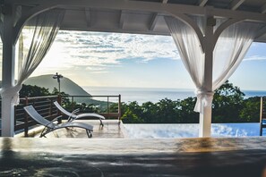 Un espace extérieur aménagé avec une zone salon couverte, des rideaux blancs, une vue sur la mer et un paysage montagneux.