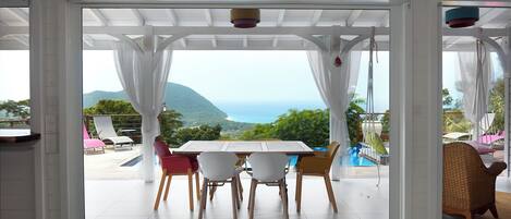 A dining area with a table and chairs, a view of the sea and mountains, and a pool in the background.