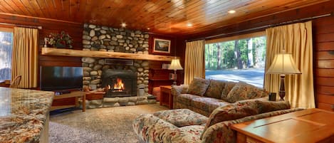 A cozy living room with a stone fireplace, wooden ceiling, and a large window with curtains.