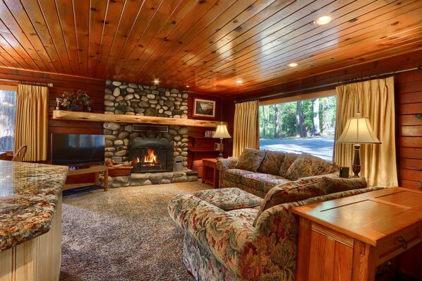 A cozy living room with a stone fireplace, wooden ceiling, and a large window with curtains.
