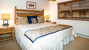 Un dormitorio con cabecera de madera, dos mesitas de noche con lámparas, una cama grande con ropa de cama estampada y una ventana con persianas.