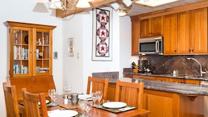 Un comedor con una mesa de madera dispuesta para cuatro personas, rodeada de sillas de madera. Hay una zona de cocina con armarios de madera y un microondas.