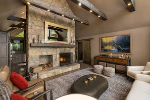 Une cheminée en pierre avec un téléviseur au-dessus, un grand tableau au mur et un coin salon confortable avec un canapé en cuir et un pouf.