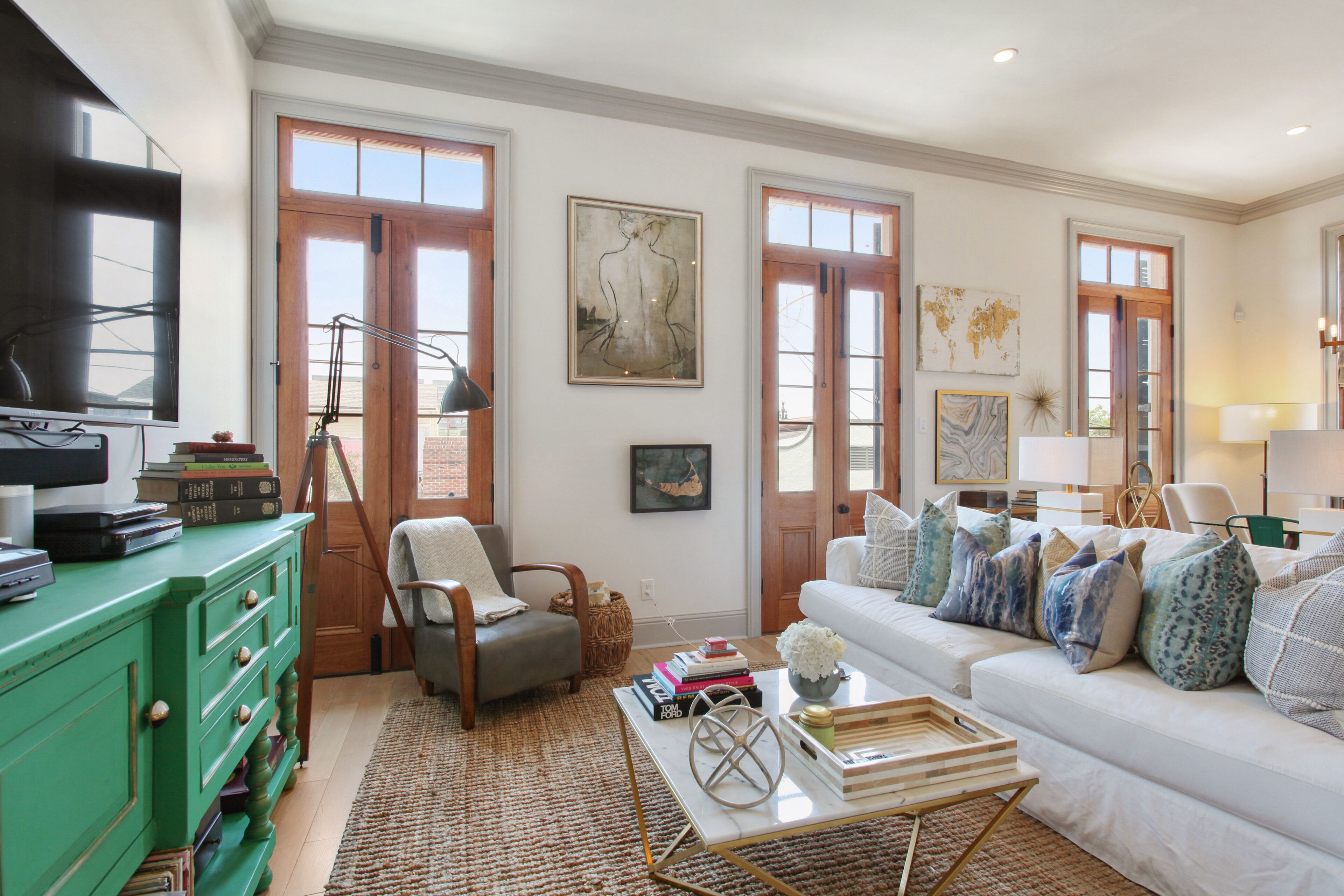 Un salon spacieux avec une commode verte, un canapé blanc orné de coussins à motifs et une table basse sur laquelle sont posés des livres et des objets décoratifs.