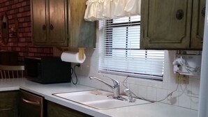 Una cocina con armarios de madera, encimera blanca, un lavamanos, microondas y una ventana con persianas.