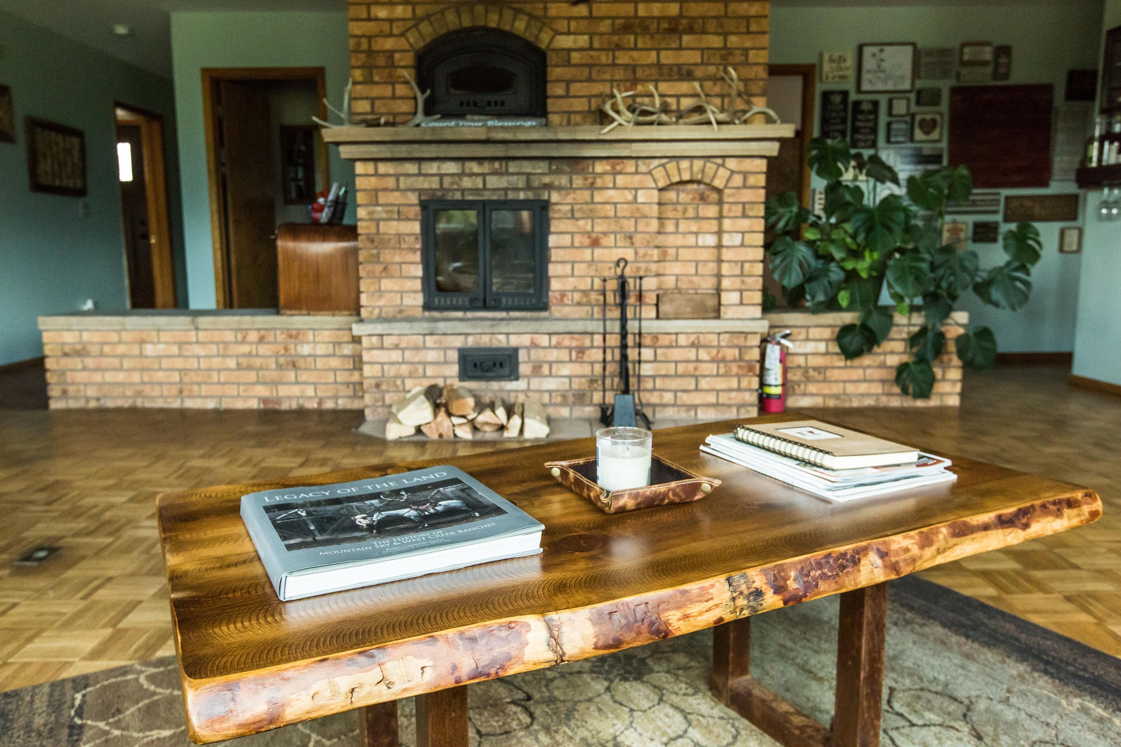 A wooden table with books and a glass of milk, a brick fireplace with a pizza oven, and a large green plant in a living room.