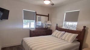 Une chambre à coucher comprenant un lit, une table de chevet, une commode, un ventilateur de plafond et un téléviseur fixé au mur.