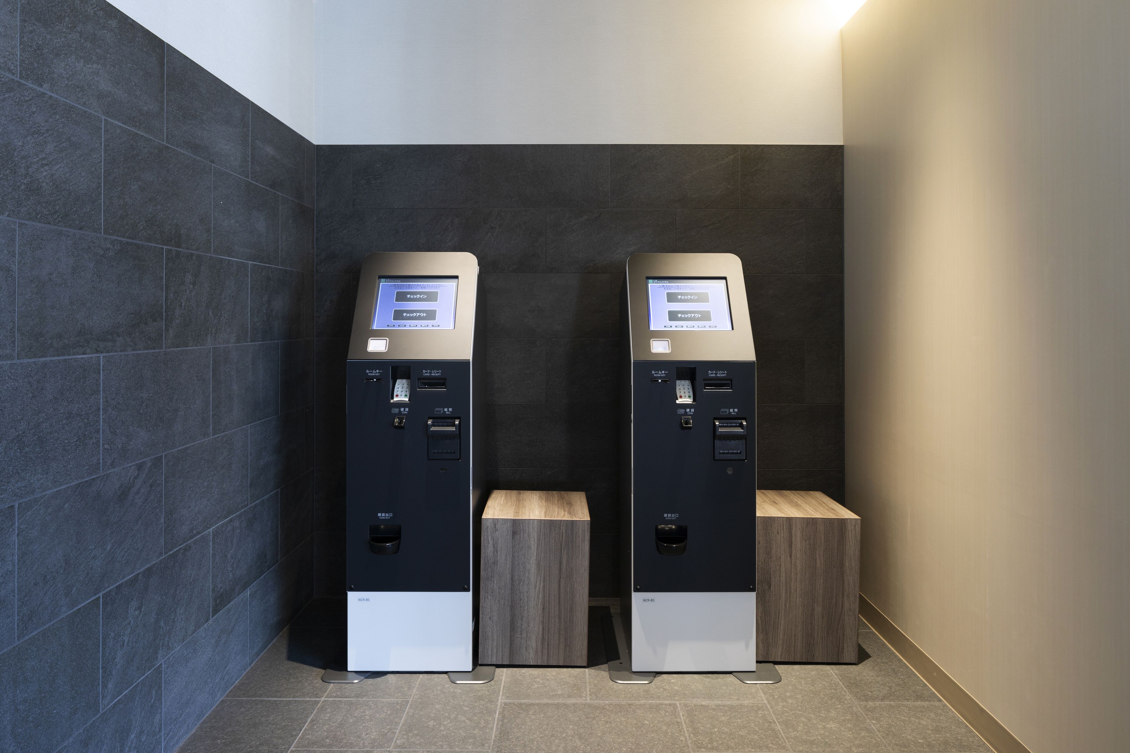 Two self-service kiosks with digital displays, placed against a dark tiled wall with a wooden accent piece in the center.