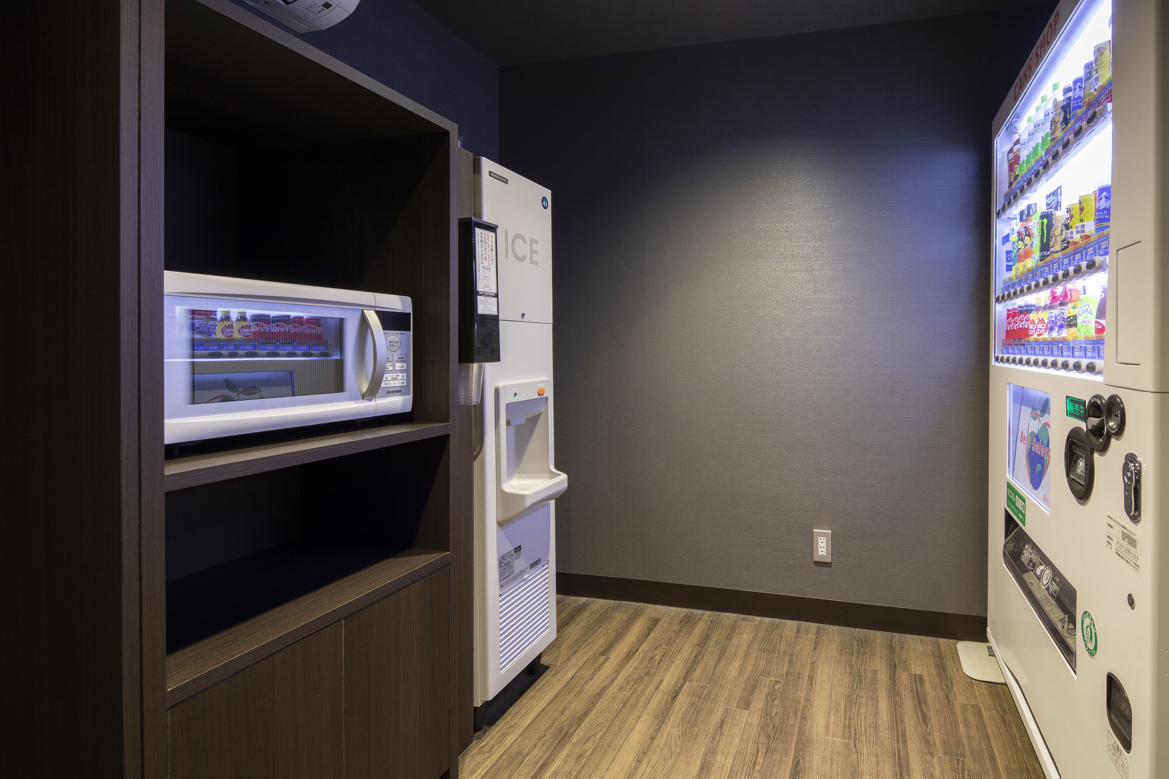Máquina expendedora con diversas bebidas y aperitivos, un microondas y un refrigerador en una habitación con suelo de madera y pared lisa.