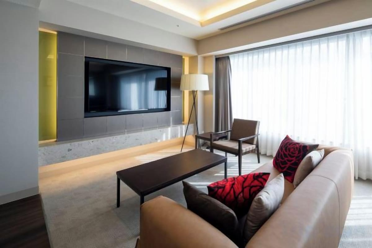 Ein modernes Hotelzimmer mit einem an der Wand befestigten Flachbildfernseher, einem Sofa mit roten und schwarzen Kissen, einem Couchtisch und einer Stehlampe.