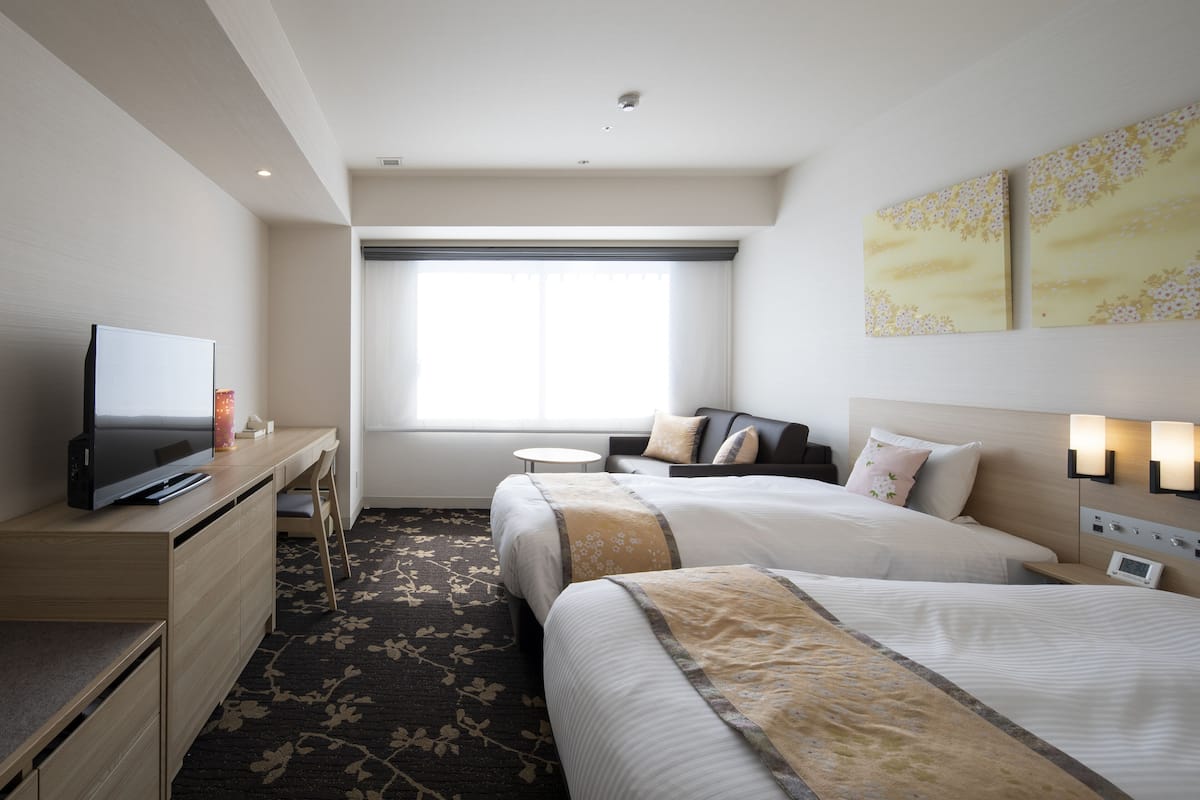 Une chambre d’hôtel avec deux lits, un bureau avec une télévision, une petite table et une fenêtre avec des rideaux.