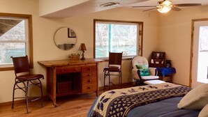 A bedroom with a wooden desk, two chairs, a bed with a patterned blanket, a ceiling fan, and large windows with a view of trees and a body of water.
