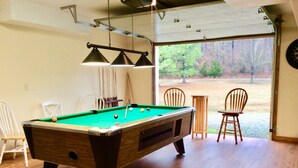 A pool table with a green felt surface, surrounded by wooden chairs, in a room with wooden flooring and a large window offering a view of trees.
