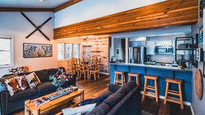 Una sala de estar moderna con un sofá, una mesa de centro y sillas de madera. Se ve una cocina en el fondo, con una encimera azul y taburetes.