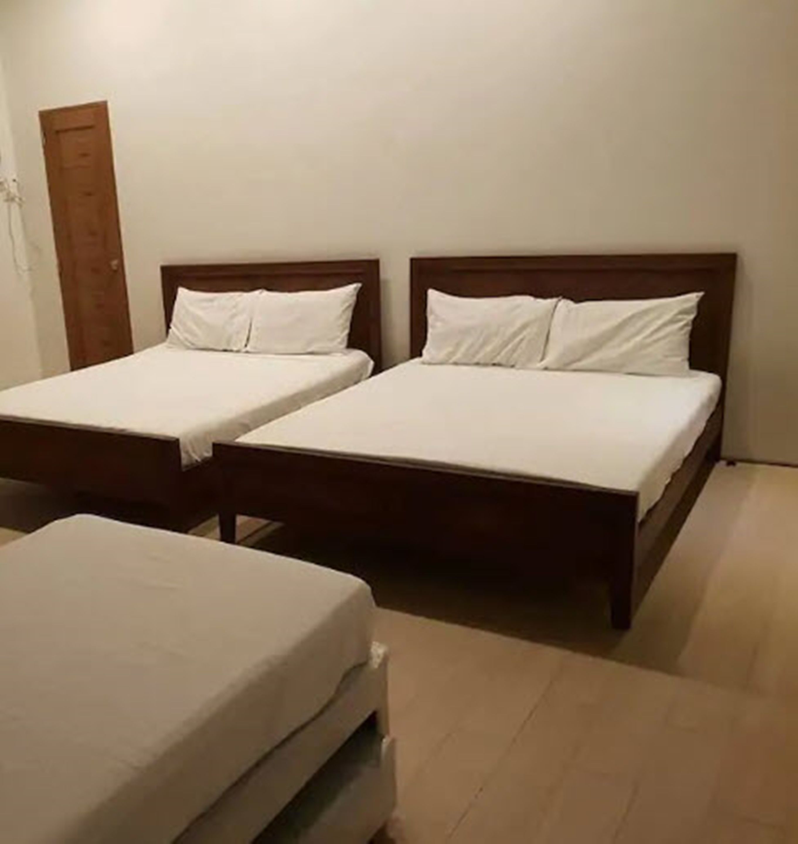 Dos camas individuales con cabeceros de madera y ropa de cama blanca en una habitación con paredes lisas y una puerta de madera.