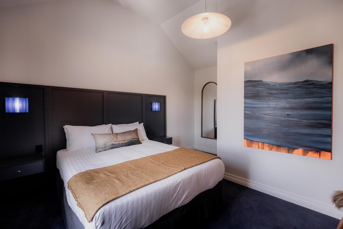 A modern bedroom with a large bed, a headboard, bedside lamps, and a painting of a seascape.