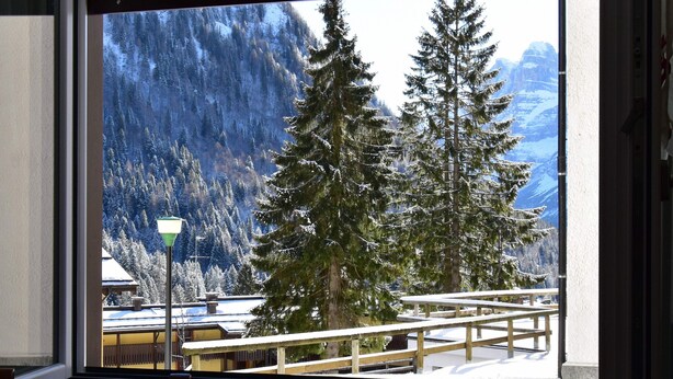 A window view of a snowy mountain landscape with tall evergreen trees and a wooden deck. - MIRA DOLO 022143-AT-347213 (Madonna di Campiglio)
