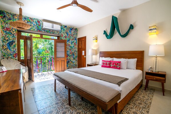 Habitación de hotel con una cama grande, cabecera de madera, dos mesitas de noche con lámparas, un banco al pie de la cama, pared colorida, puerta que da a un balcón y un ventilador de techo.