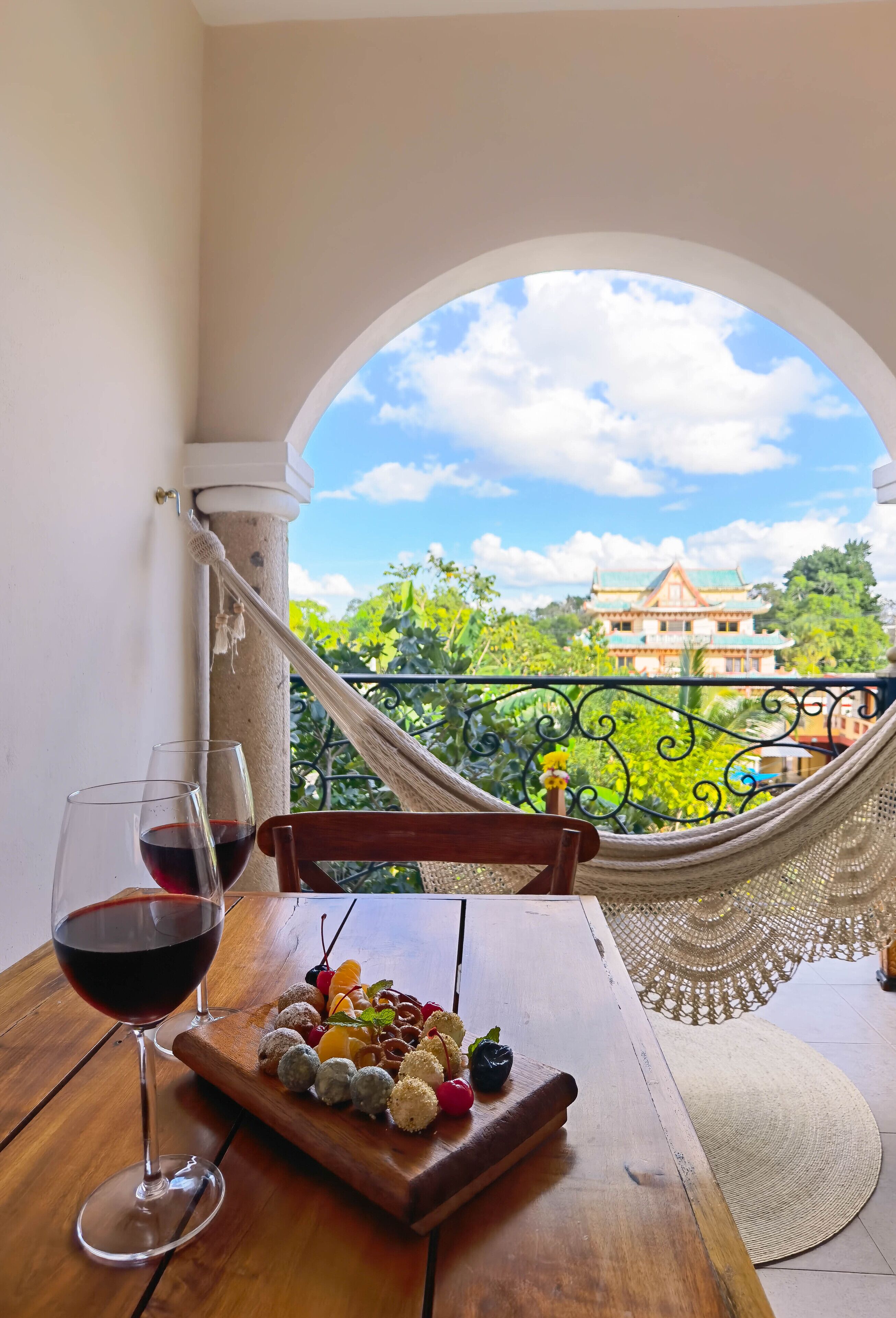 Une table en bois avec un verre de vin rouge et un plateau rempli de diverses collations, un hamac et une vue sur un bâtiment traditionnel, visible à travers une fenêtre en arc de cercle.